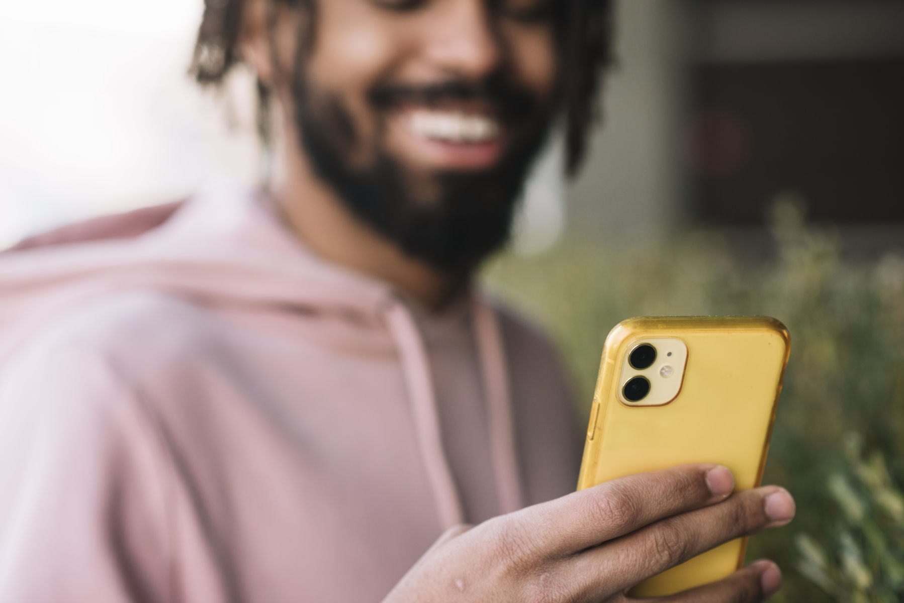 Smiling person in a pink hoodie using a smartphone with a yellow case outdoors.